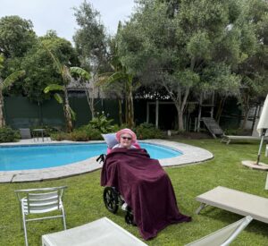 Person in a wheelchair covered with a blanket sits by a pool in a garden area with lounge chairs.